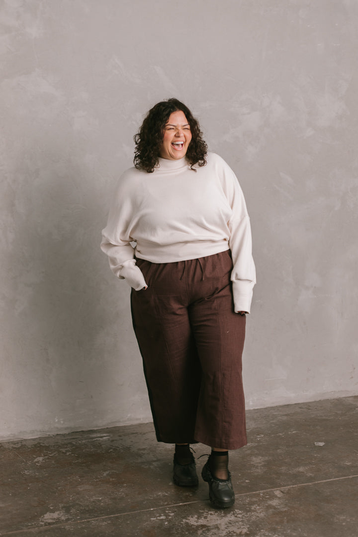 Person wearing a white sweater and brown pants against a gray wall.